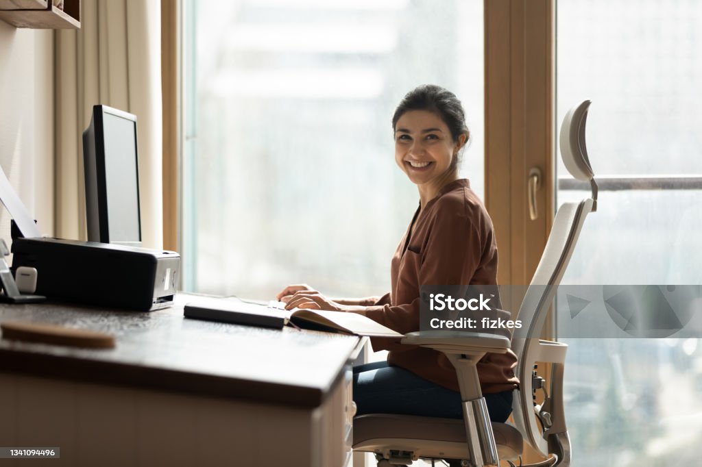 Modern day worker. Portrait of happy biracial business woman freelancer sit by computer at comfy workplace at corporate workspace or at home. Smiling young indian lady office employee look at camera